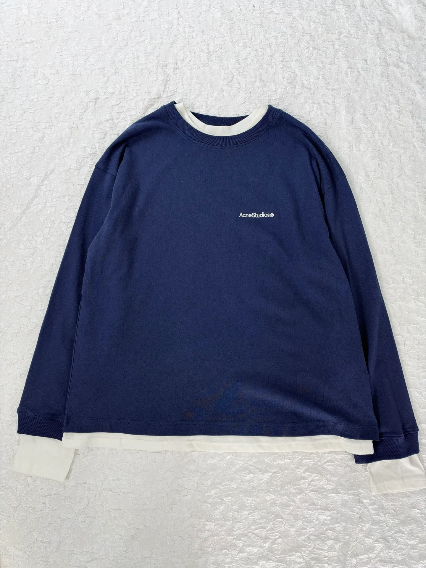 Acne Studio letter logo embroidered fake two-piece deconstructed long-sleeved T-shirt. Made of 100% cotton knitted plain fabric. The garment is pre-shrunk and washed to effectively reduce washing shrinkage. The version adopts dropped shoulder sleeves + loose box cut, the round neck is equipped with a rib collar, and the binding strip is used under the collar for reinforcement. The front chest uses high-density computer embroidery technology to present the brand logo. Visually, the inner and outer layers are designed with contrasting color splicing. The hem and cuffs are double-layered fake two-piece stacking. The sewing adopts flat seam + overlock technology. The lines are even and dense, and the overall relaxed silhouette takes into account practical wearability and layering effect. It can be worn alone or layered. Size：S/M/L/XL
